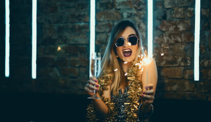 Woman wearing a sparkly dress and holding an alcoholic beverage and sparkler in her hands.