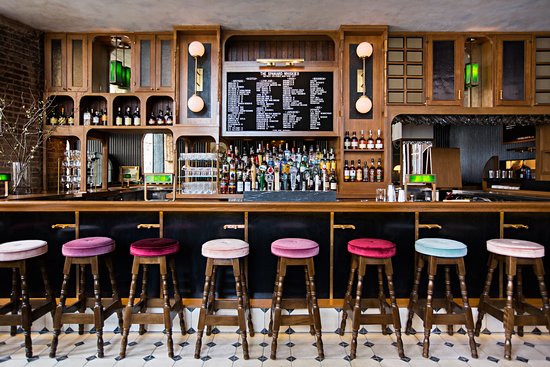 Black and white floor tiles and velvet topped bar stools at a wood-covered bar.