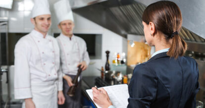 A woman who is the restaurant general manager is taking notes while talking to two chefs wearing white uniforms in a commercial kitchen.