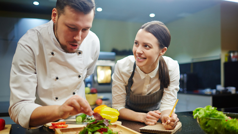 Young woman writing down recipe of vegetable salad while chief cooking it