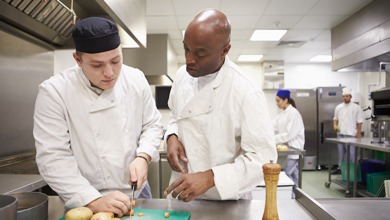 Teacher Helping Students Training To Work In Catering Chopping Vegetables