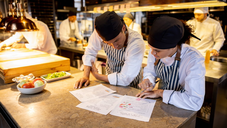 Two chefs working in a restaurant kitchen looking at a recipe for a menu item.