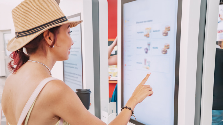 Woman wearing a beige hat ordering food from a self ordering kiosk.