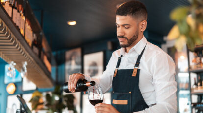 Man in a white dress shirt and black apron standing pouring a glass of red wine.